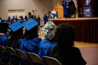Graduates in blue robes sit facing a stage. One cap is decorated with blue and white flowers, and a message