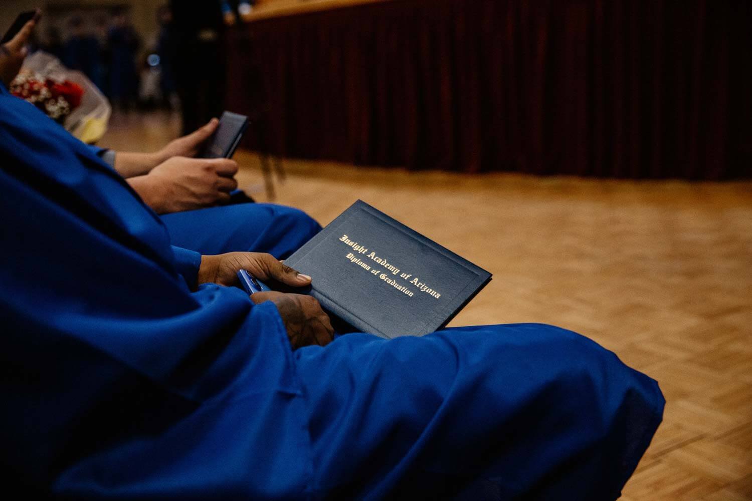 A graduate in a blue gown holds a diploma at a graduation ceremony.