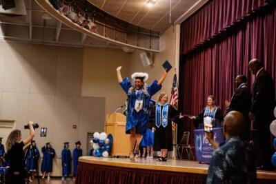 Graduate in a blue robe joyfully raises arms and cap on stage.