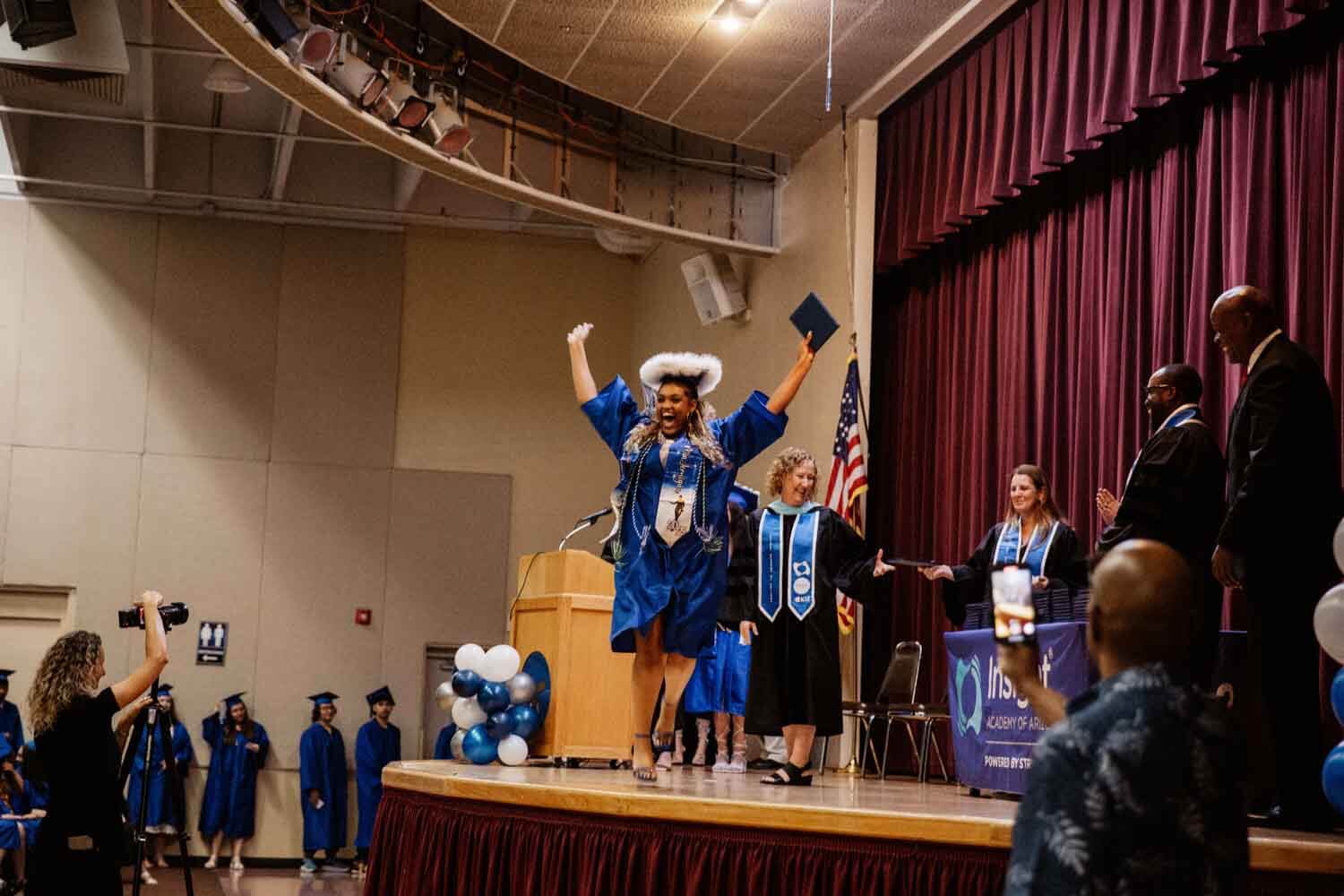 Graduate in a blue robe joyfully raises arms and cap on stage.