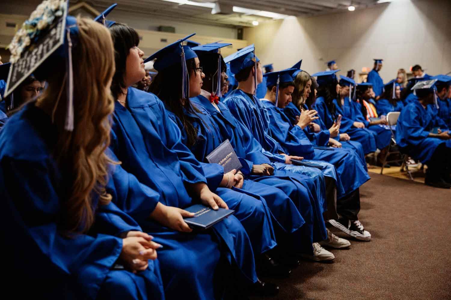 Graduates in blue caps and gowns sit in rows at a ceremony.