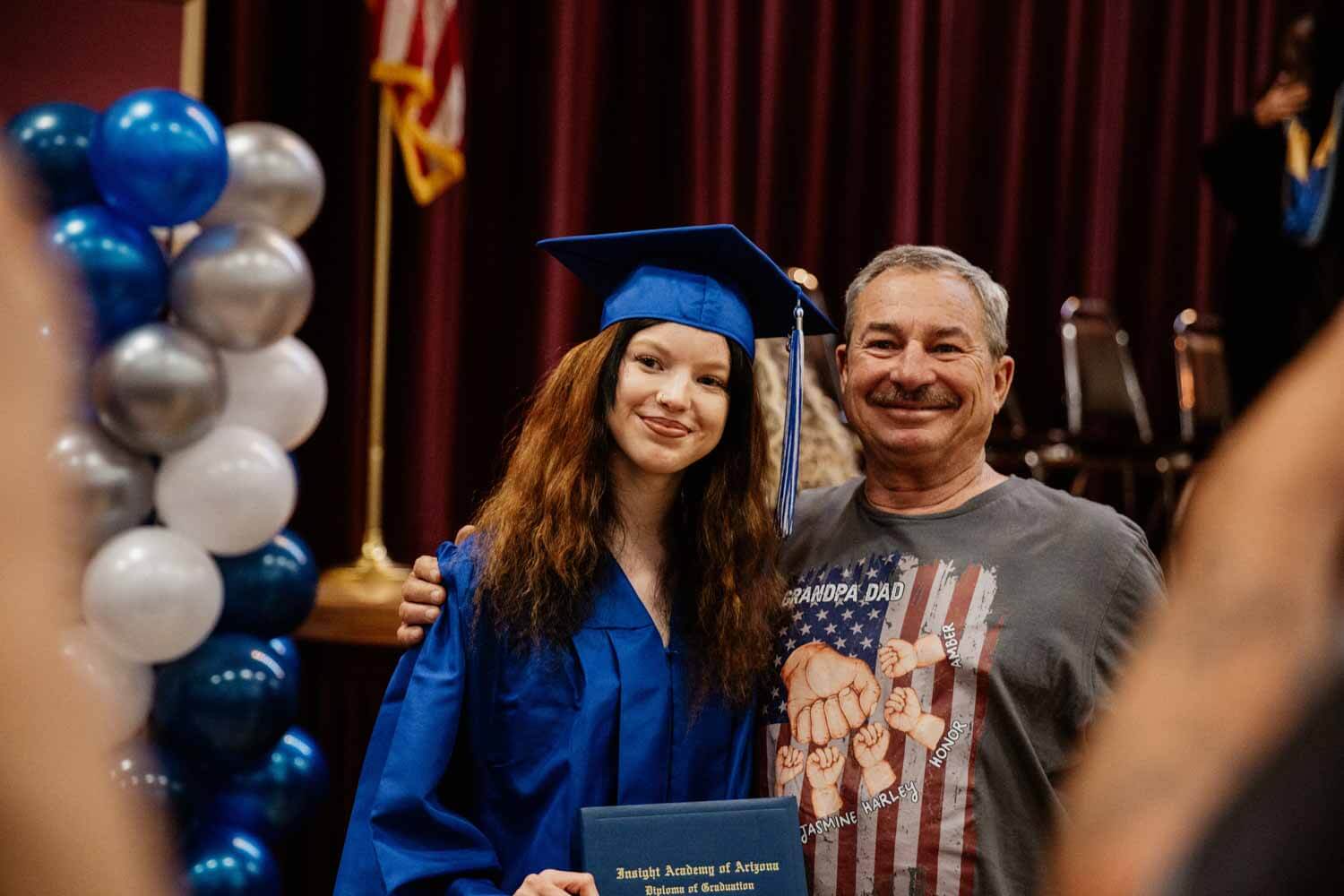 A graduate in a blue cap and gown smiles beside a proud older man.