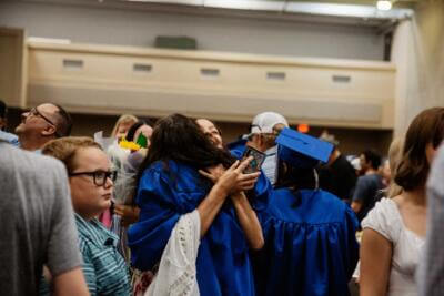 Graduates in blue gowns and caps embrace joyfully in a crowded indoor setting.