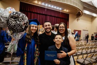 Graduate in blue cap and gown holds "Class of 2025" balloons, standing with smiling family in a hall.