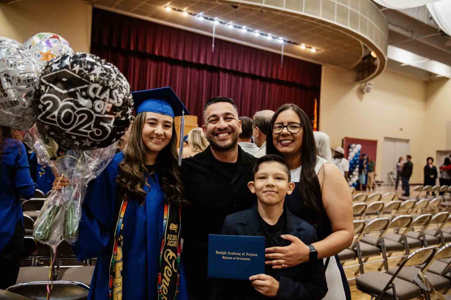 Graduate in blue cap and gown holds "Class of 2025" balloons, standing with smiling family in a hall.
