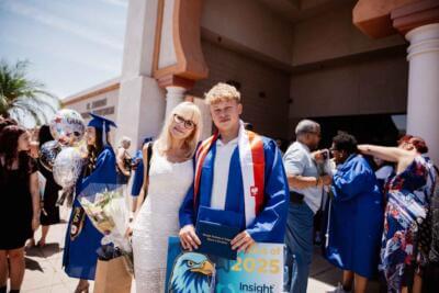 A young graduate in a blue gown with red trim stands with a woman in a white dress, holding a "Class of 2025" sign.