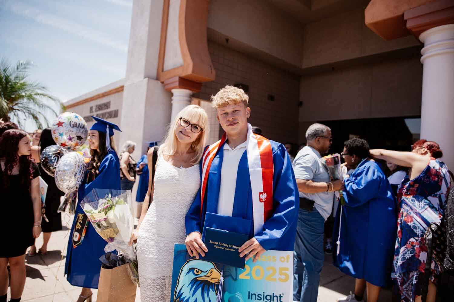A young graduate in a blue gown with red trim stands with a woman in a white dress, holding a "Class of 2025" sign.