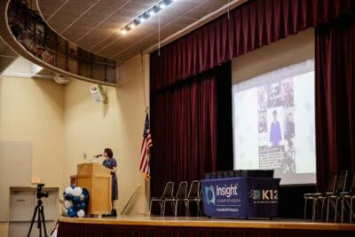A woman speaks at a podium on a school stage with an American flag