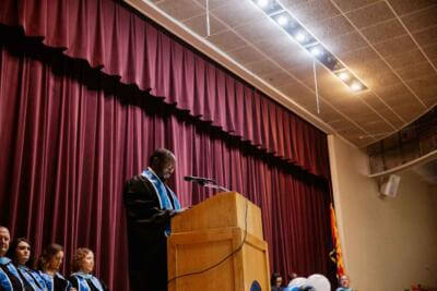 A speaker in a graduation robe stands at a podium on a stage with a maroon curtain.