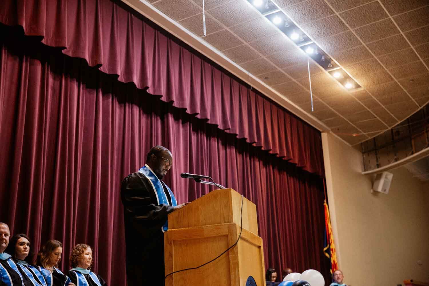 A speaker in a graduation robe stands at a podium on a stage with a maroon curtain.