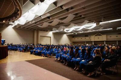 A graduation ceremony in a spacious hall with graduates in blue robes seated in rows.