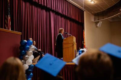 A person in academic attire speaks at a wooden podium on a stage with maroon curtains