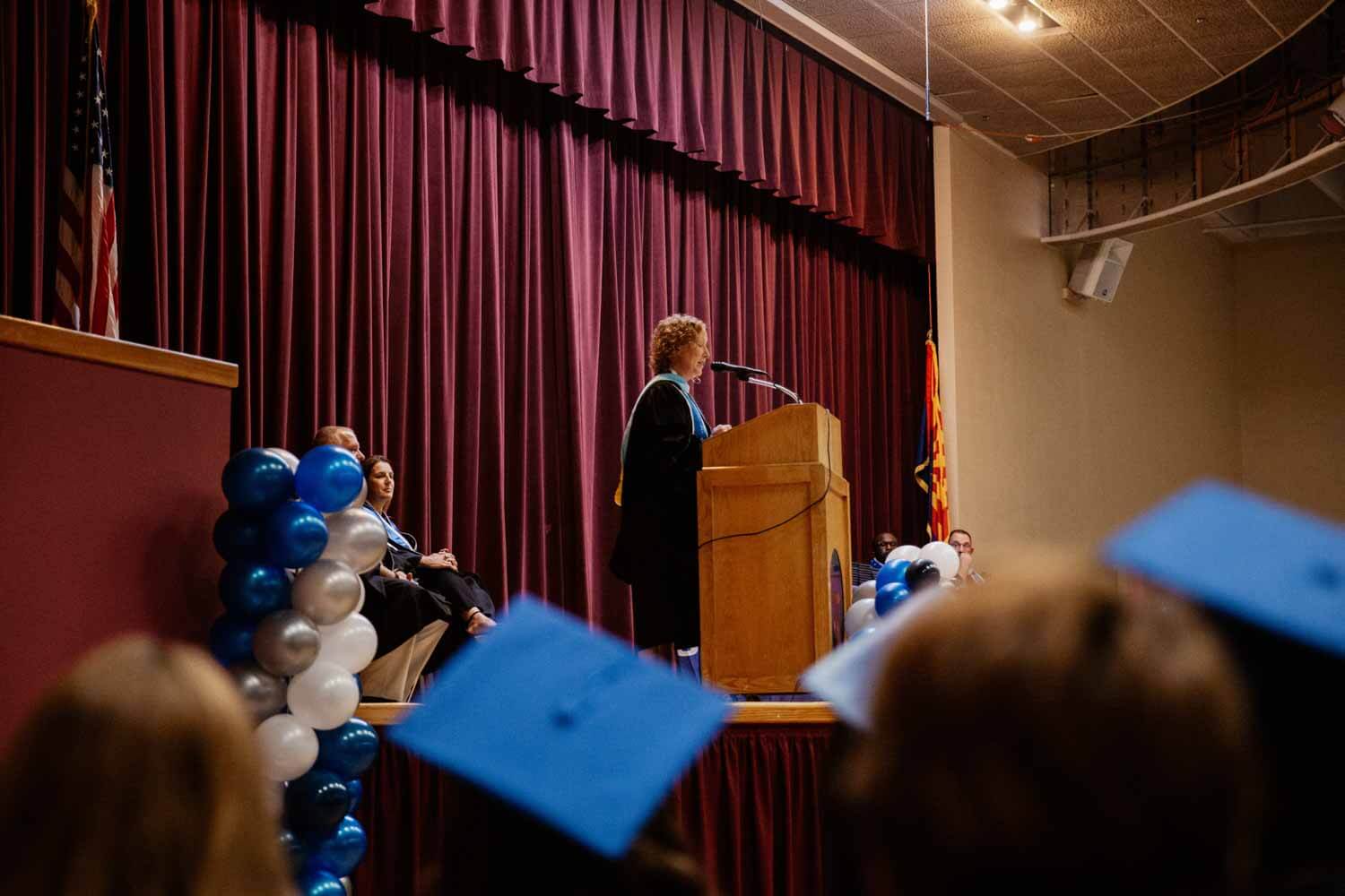 A person in academic attire speaks at a wooden podium on a stage with maroon curtains