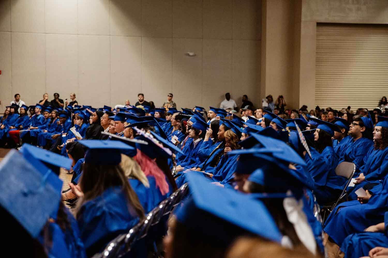 Graduates in blue caps and gowns sit in rows during a ceremony.
