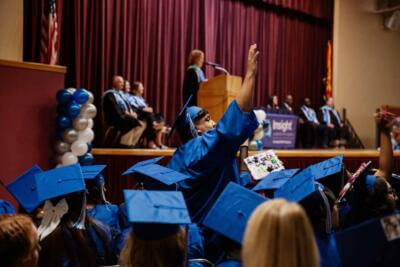 Graduates in blue caps and gowns sit excitedly as one raises their hand during a graduation ceremony.