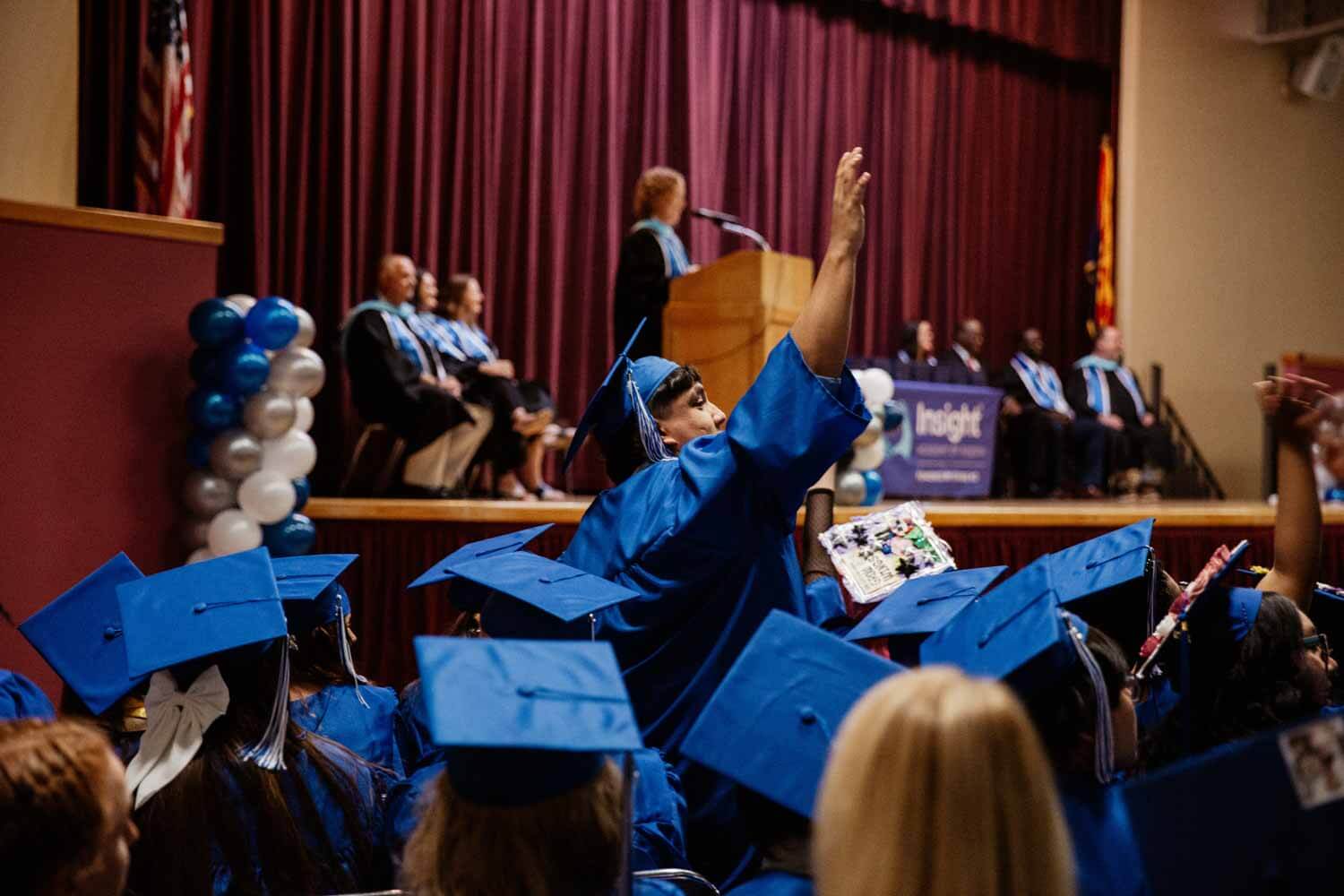 Graduates in blue caps and gowns sit excitedly as one raises their hand during a graduation ceremony.