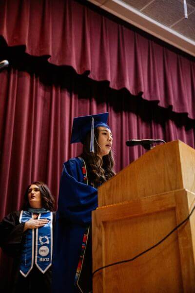 A graduate in a blue cap and gown speaks at a podium onstage, conveying confidence.