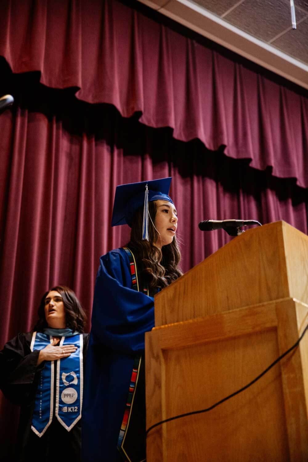 A graduate in a blue cap and gown speaks at a podium onstage, conveying confidence.