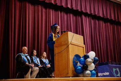 A graduate in a blue cap and gown delivers a speech at a podium on stage.