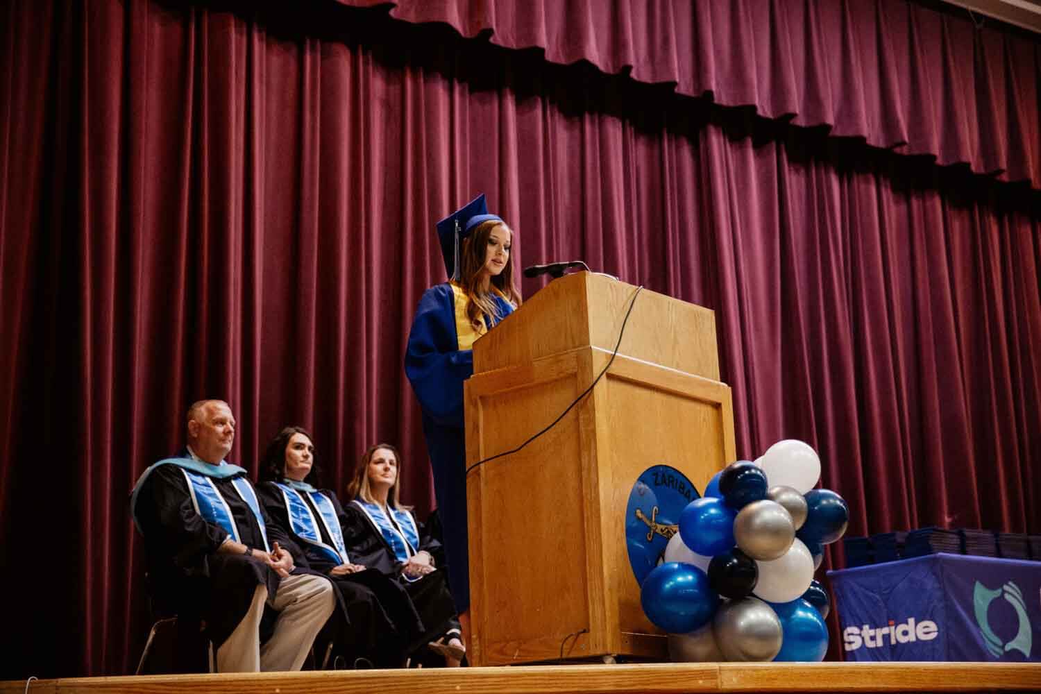 A graduate in a blue cap and gown delivers a speech at a podium on stage.