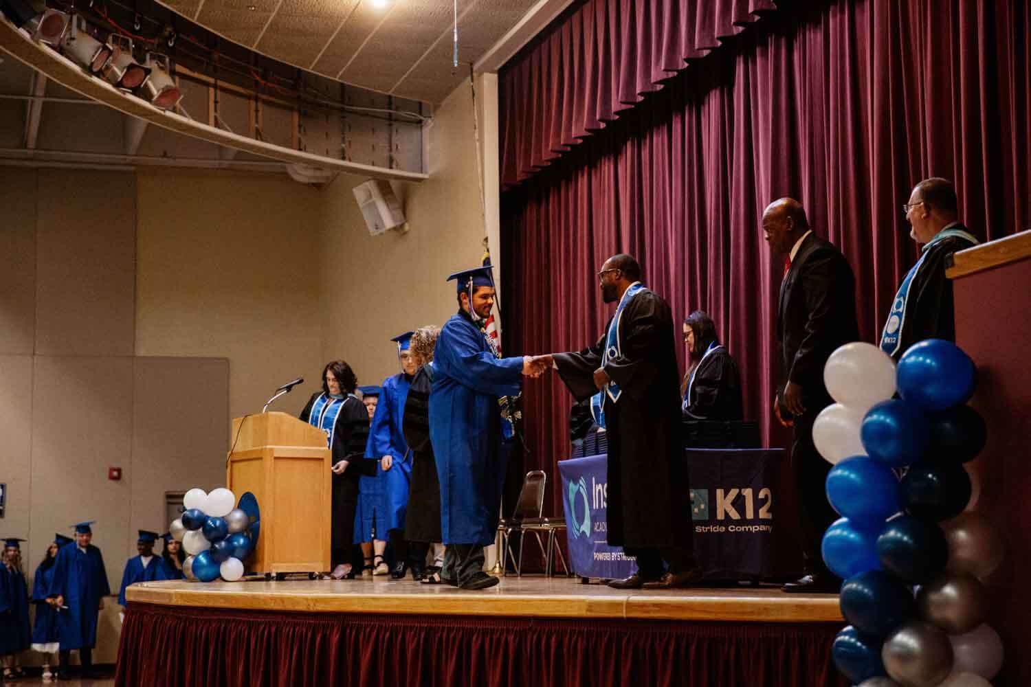 A graduate in a blue cap and gown shakes hands with a faculty member on a stage during a ceremony.