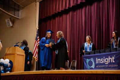 Graduation ceremony with a student in a blue cap and gown receiving a diploma on stage.