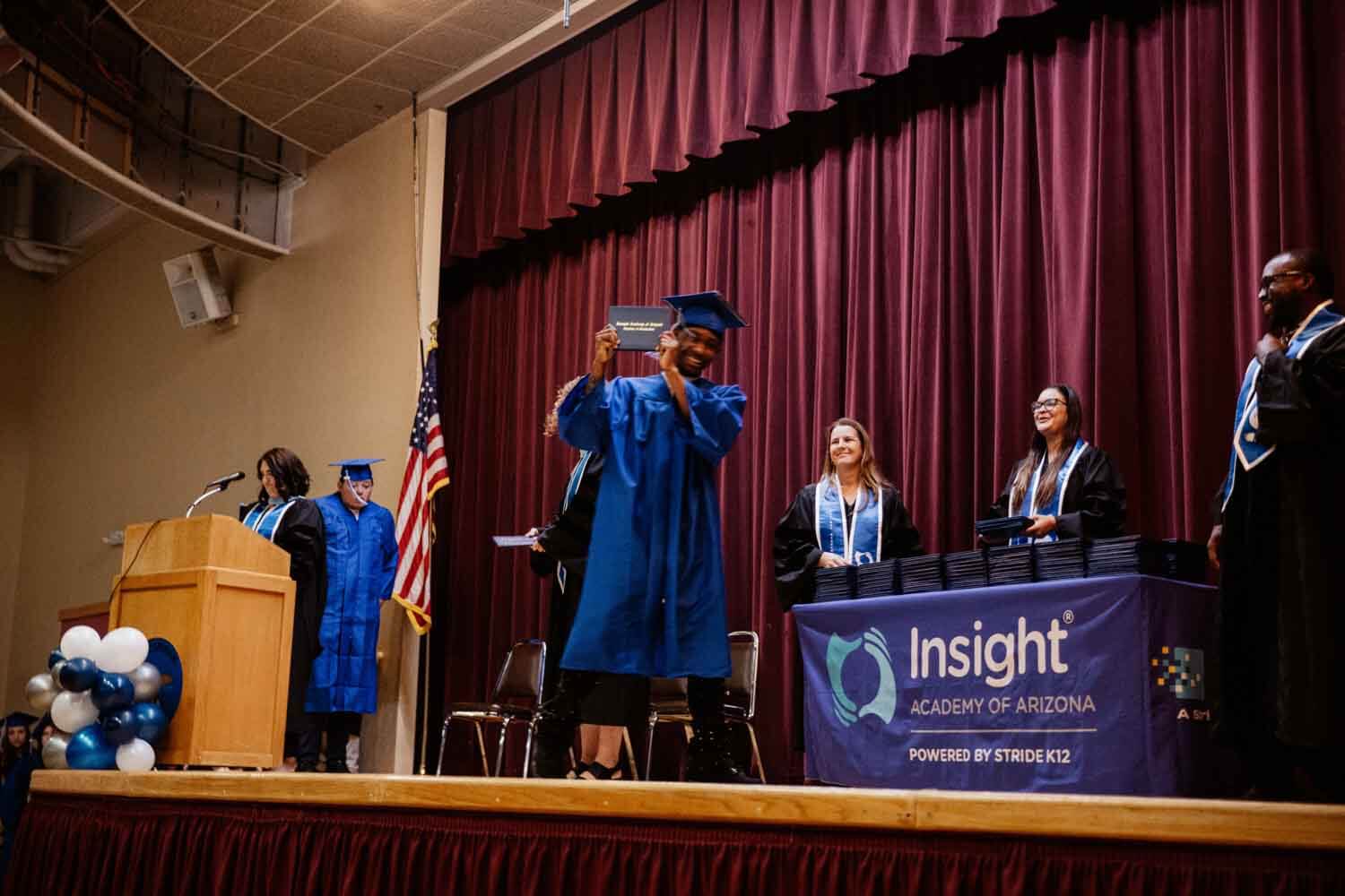 A graduate in a blue cap and gown joyfully holds up a diploma on stage