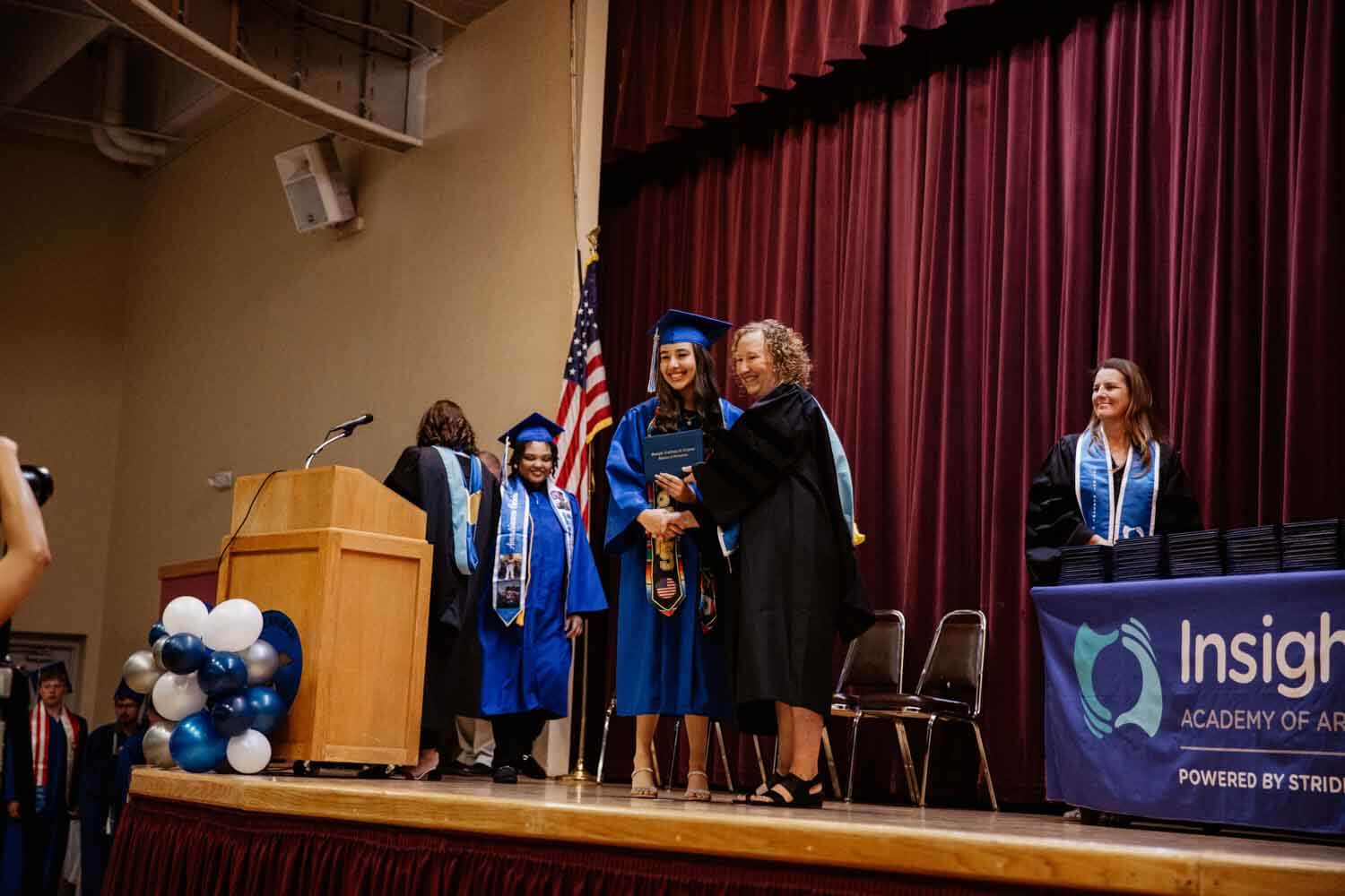 Graduation ceremony with a smiling graduate in blue robes receiving a diploma on stage.